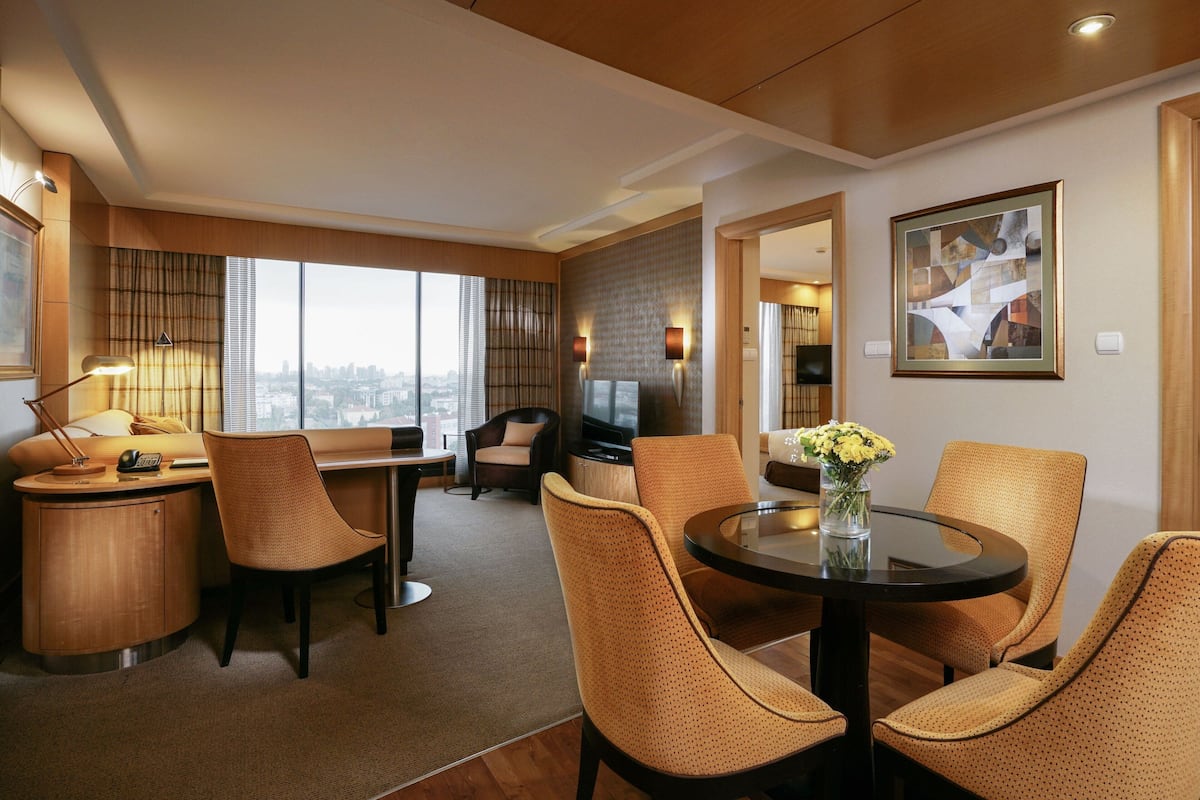 A modern hotel room with a dining area, a desk, and a large window offering a city view.