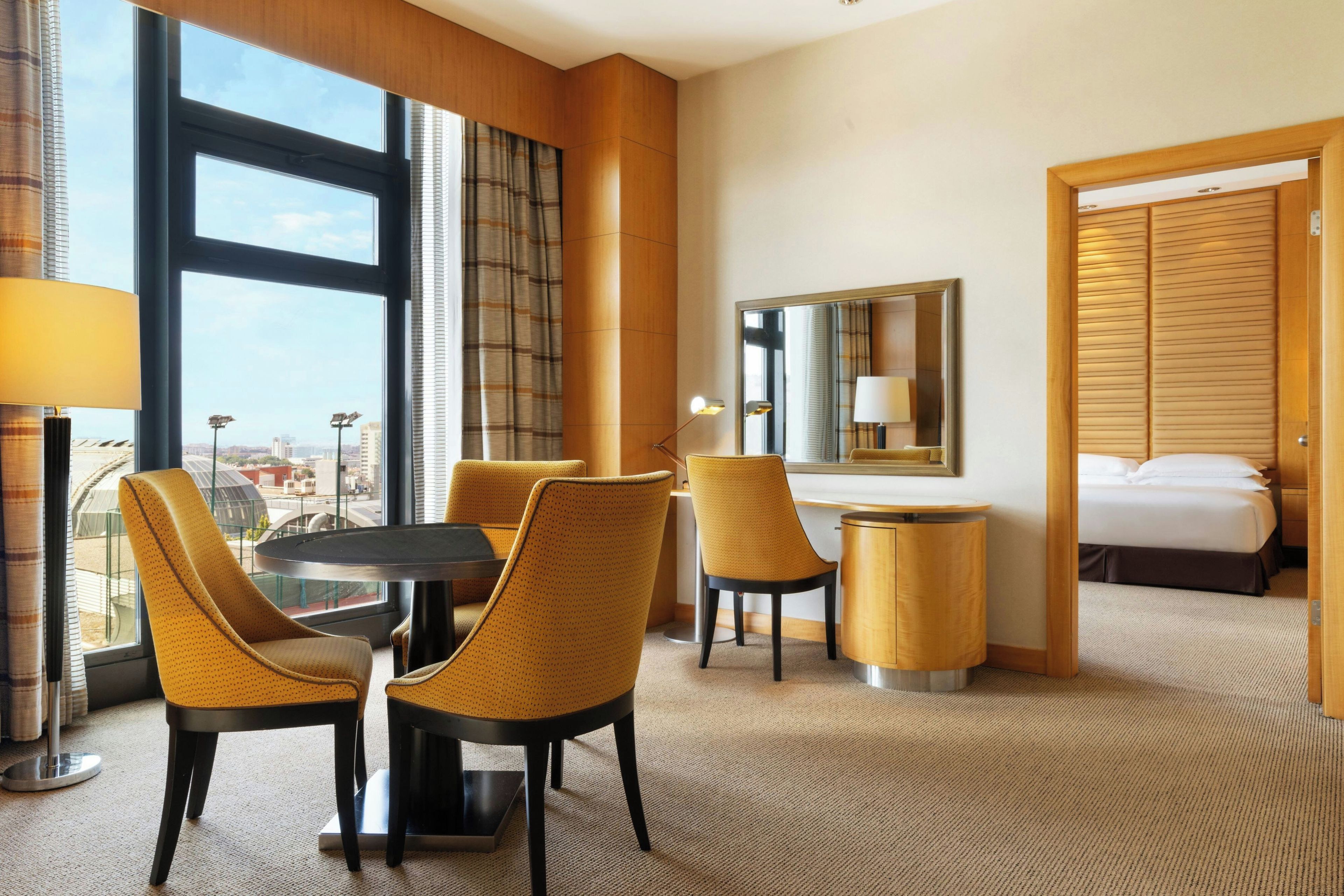 A hotel room with a large window, a round table, two chairs, a bed, a bedside table, a lamp, and a mirror.