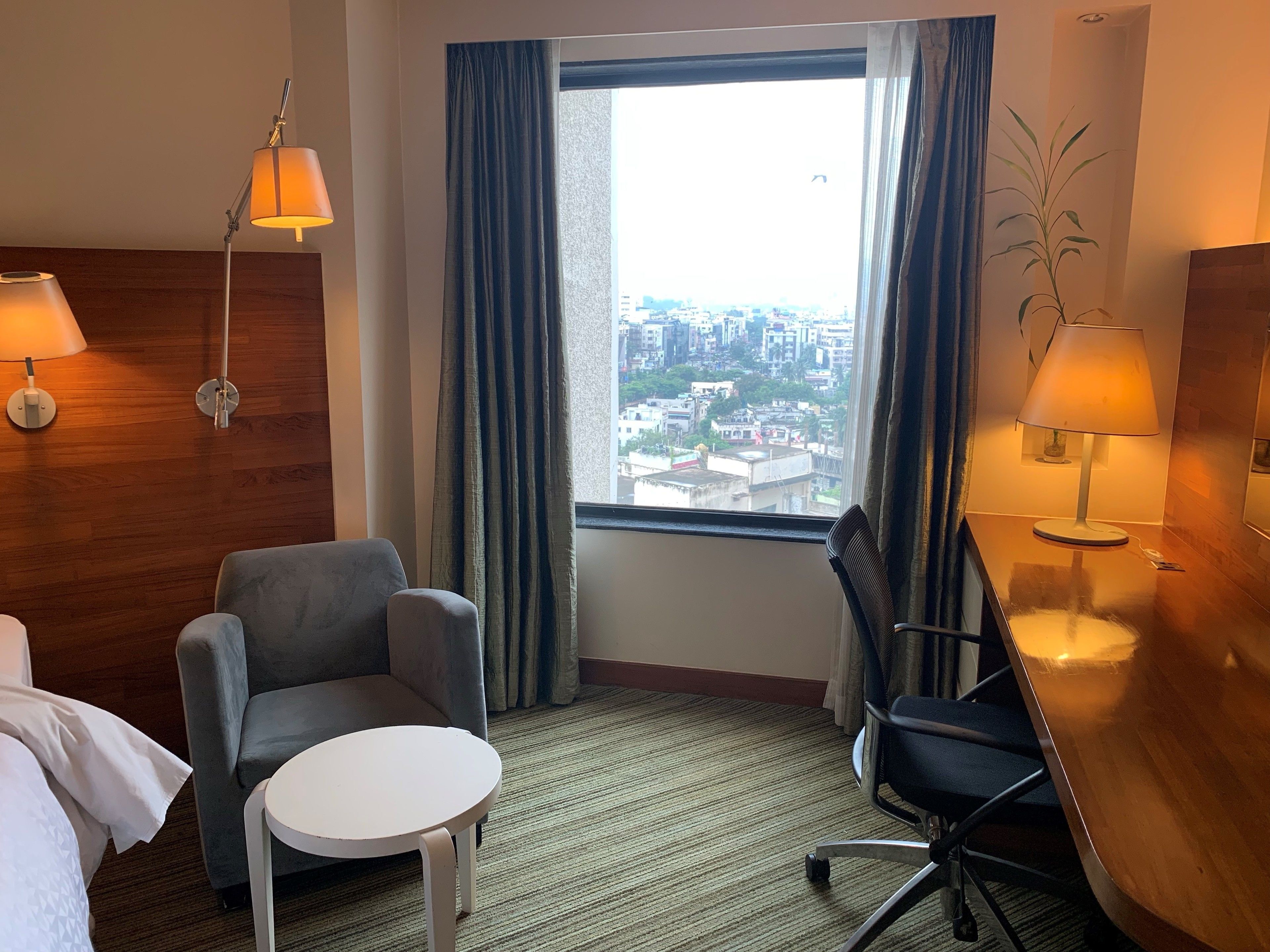 Habitación de hotel con vista a un paisaje urbano a través de un ventanal grande, un sillón gris, una mesita blanca pequeña y dos lámparas a cada lado de la ventana.