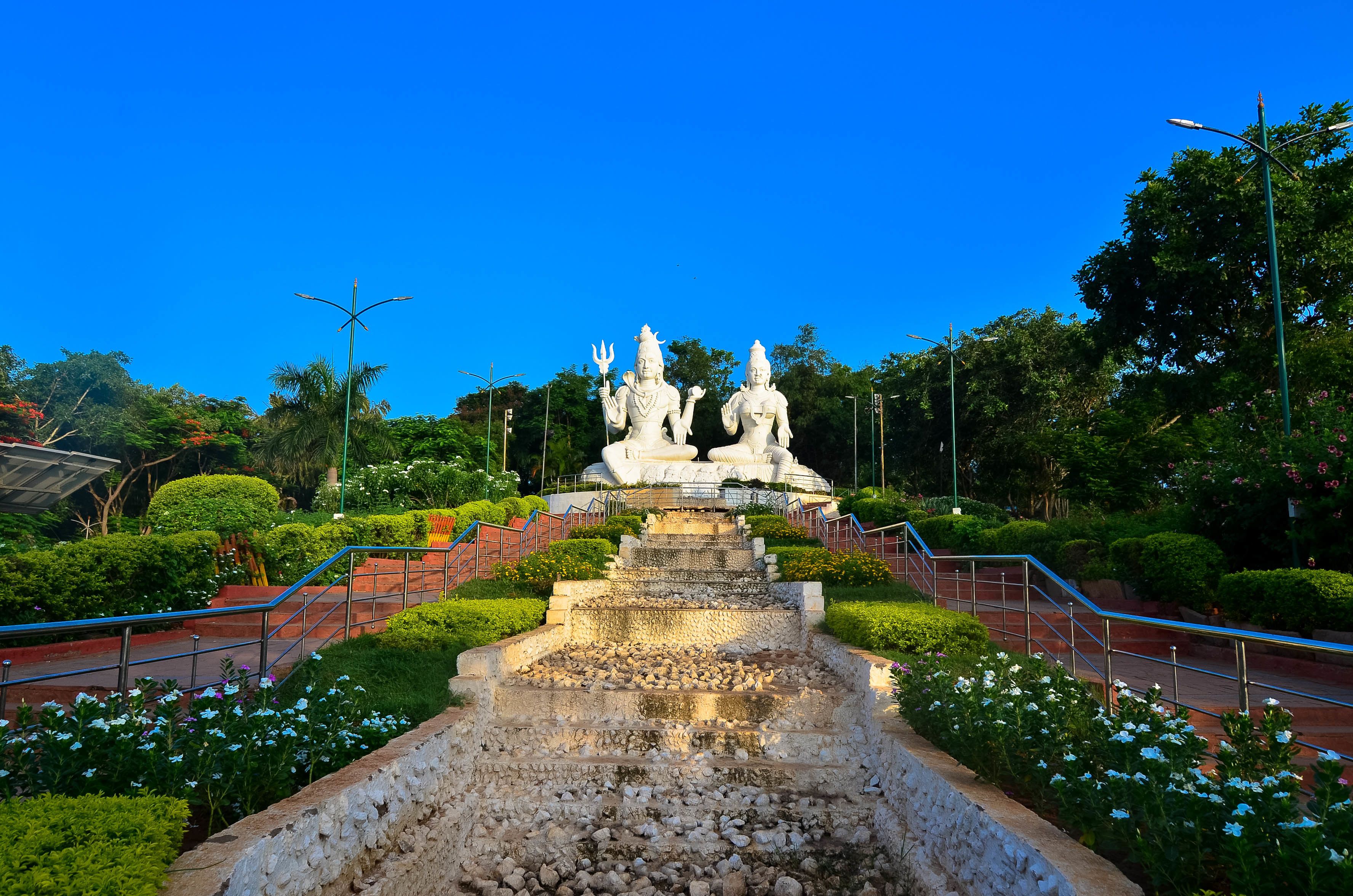 A temple with a stone staircase leading up to a statue, surrounded by greenery and a clear blue sky.