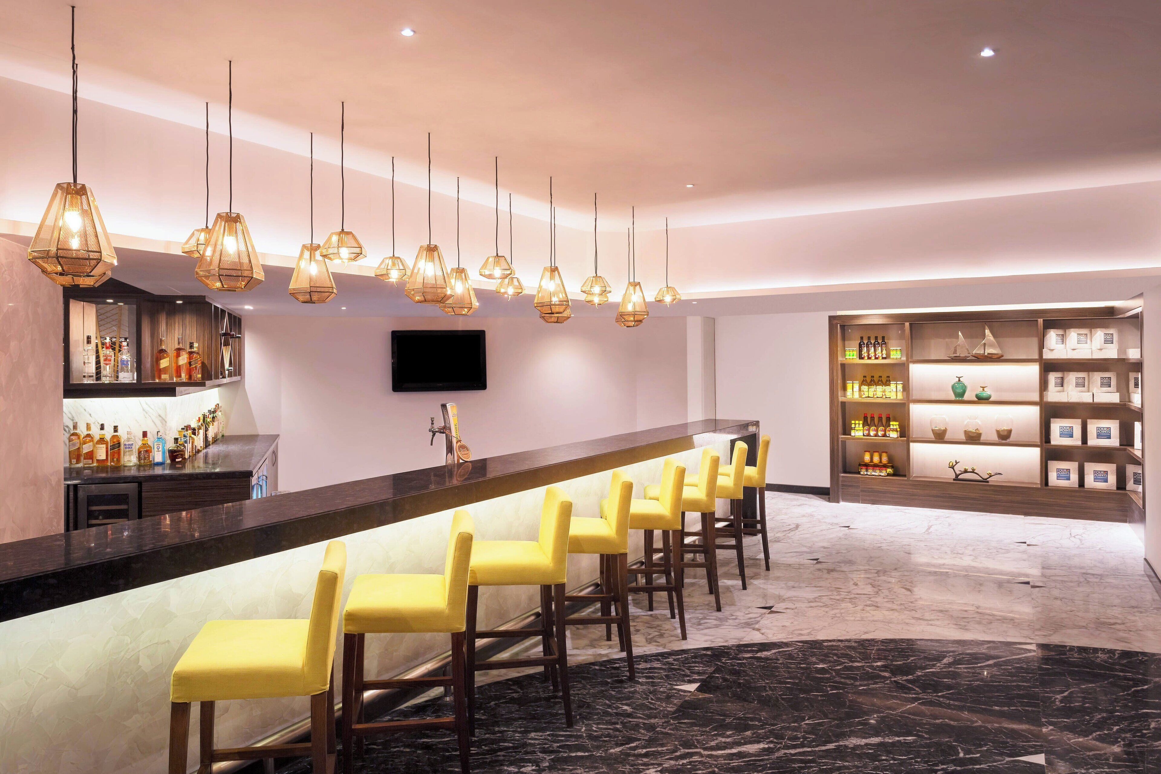 Zona de bar con una barra larga, varios taburetes amarillos, lámparas colgantes y estantes con botellas y objetos decorativos.
