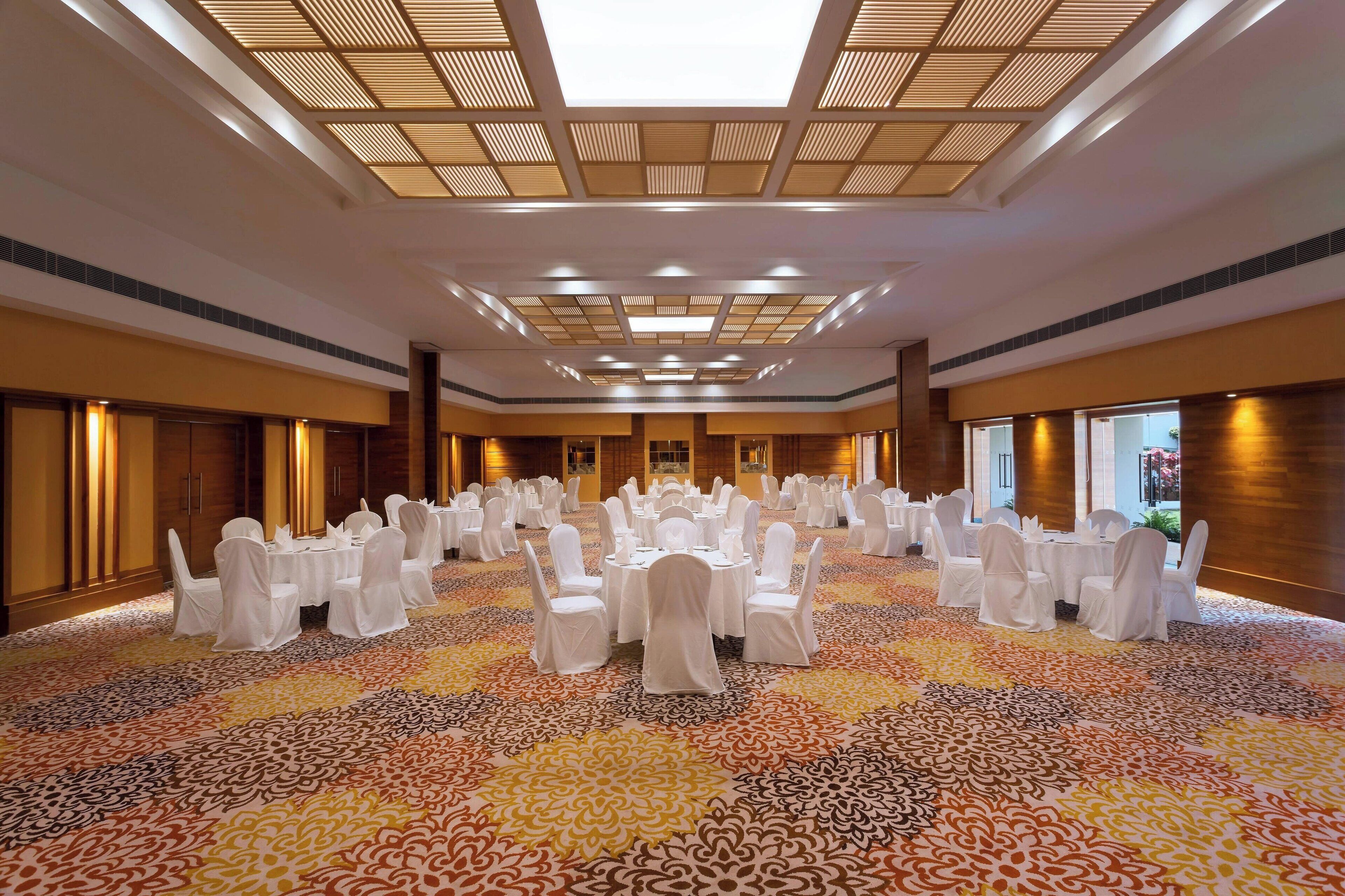 Une vaste salle de banquet avec des tables rondes et des chaises blanches, une moquette à motifs et un plafond haut doté de verrières.