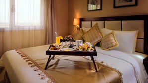 Habitación de hotel con una cama bien hecha, una bandeja con alimentos para el desayuno, una lámpara de noche y cuadros en la pared.