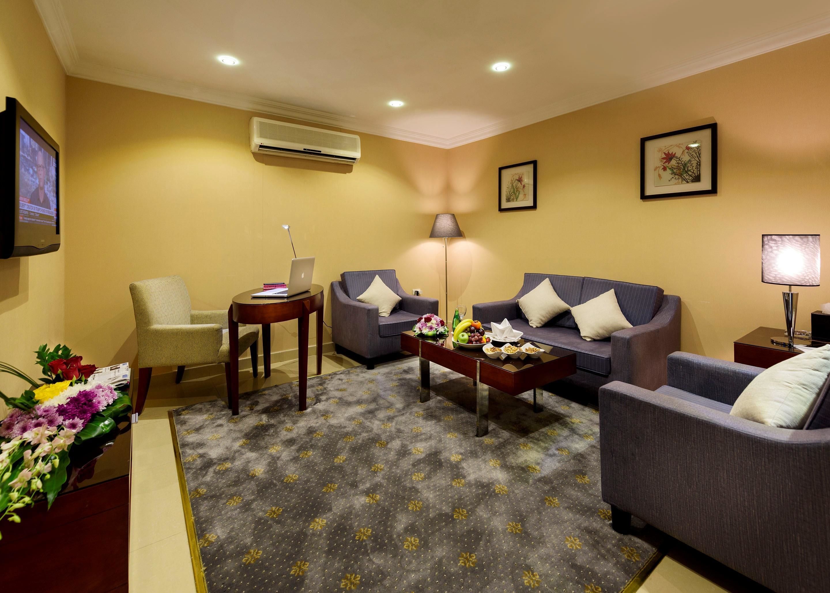 Habitación de hotel con sofá, sillones, una mesita de centro, televisor, aire acondicionado y un arreglo floral.