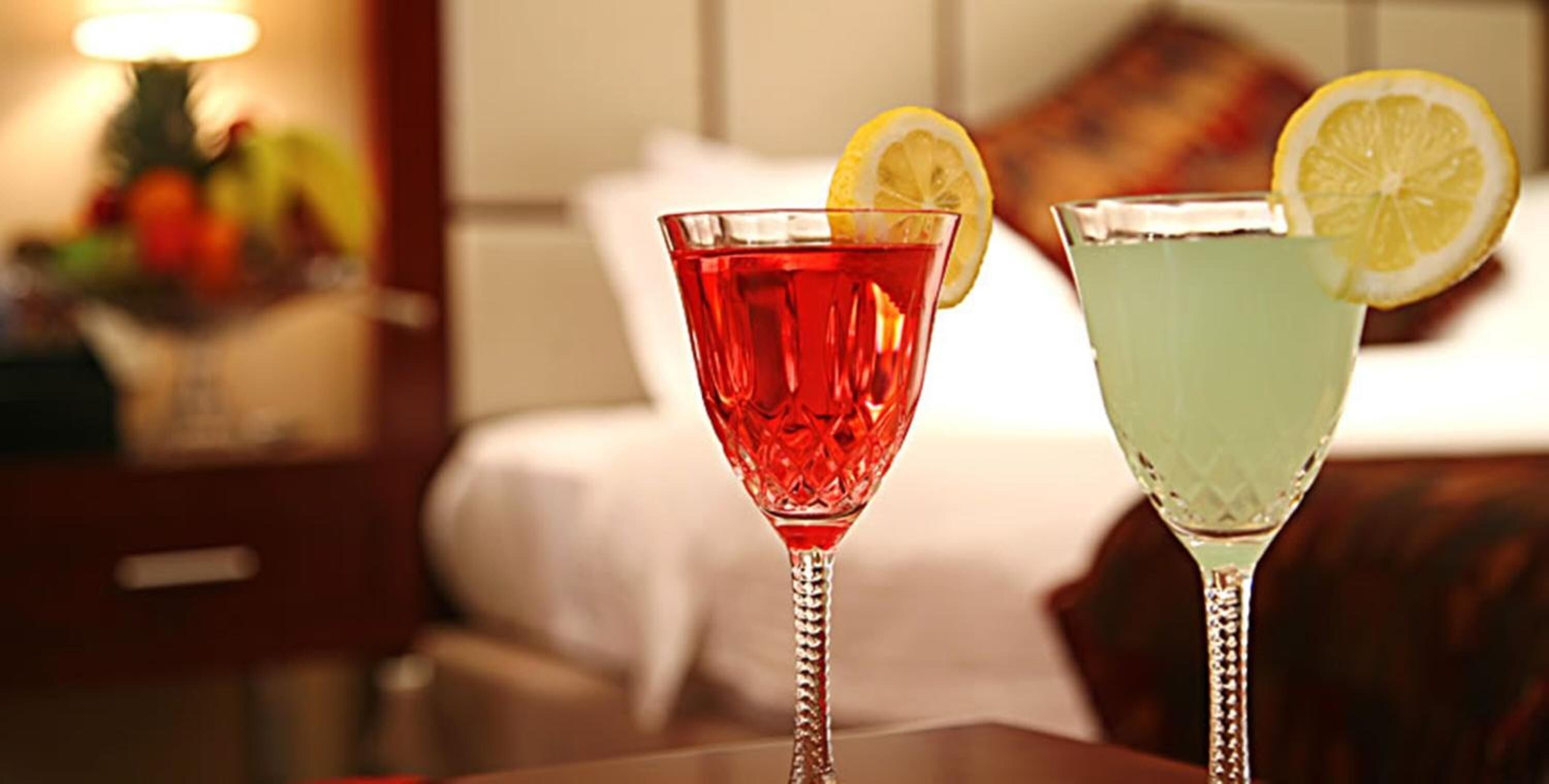 Duas taças de cocktail sobre uma mesa, com um fundo desfocado de uma cama e uma mesinha de cabeceira.