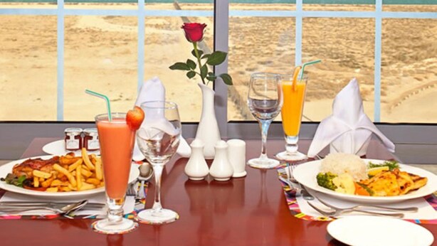 Mesa para dos con vista al mar a través de amplios ventanales, una rosa sobre la mesa y una variedad de bebidas y platos.