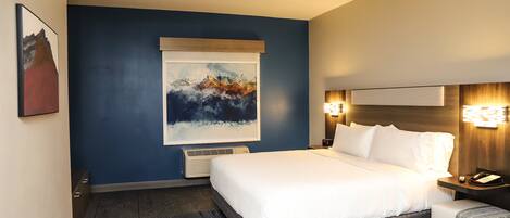 A modern hotel room with a large bed, a bedside table with a lamp, a wall-mounted air conditioner, and a framed abstract painting.