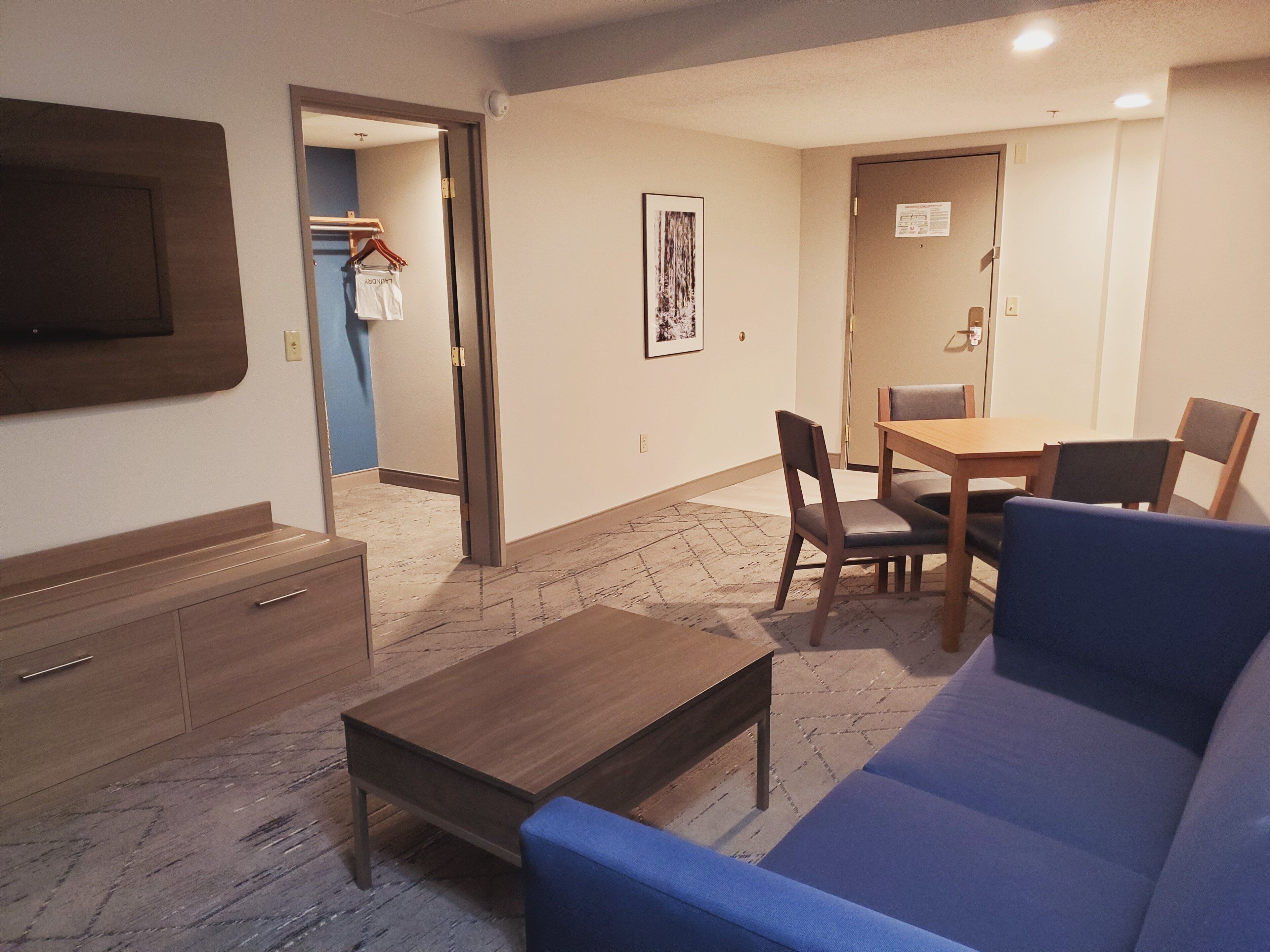 A hotel room with a sofa, a wooden table, a TV mounted on the wall, and a door leading to another room.