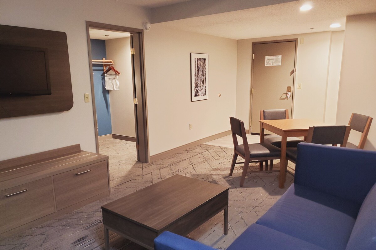 A room with a sofa, a wooden table, a TV mounted on the wall, and a door leading to another room.