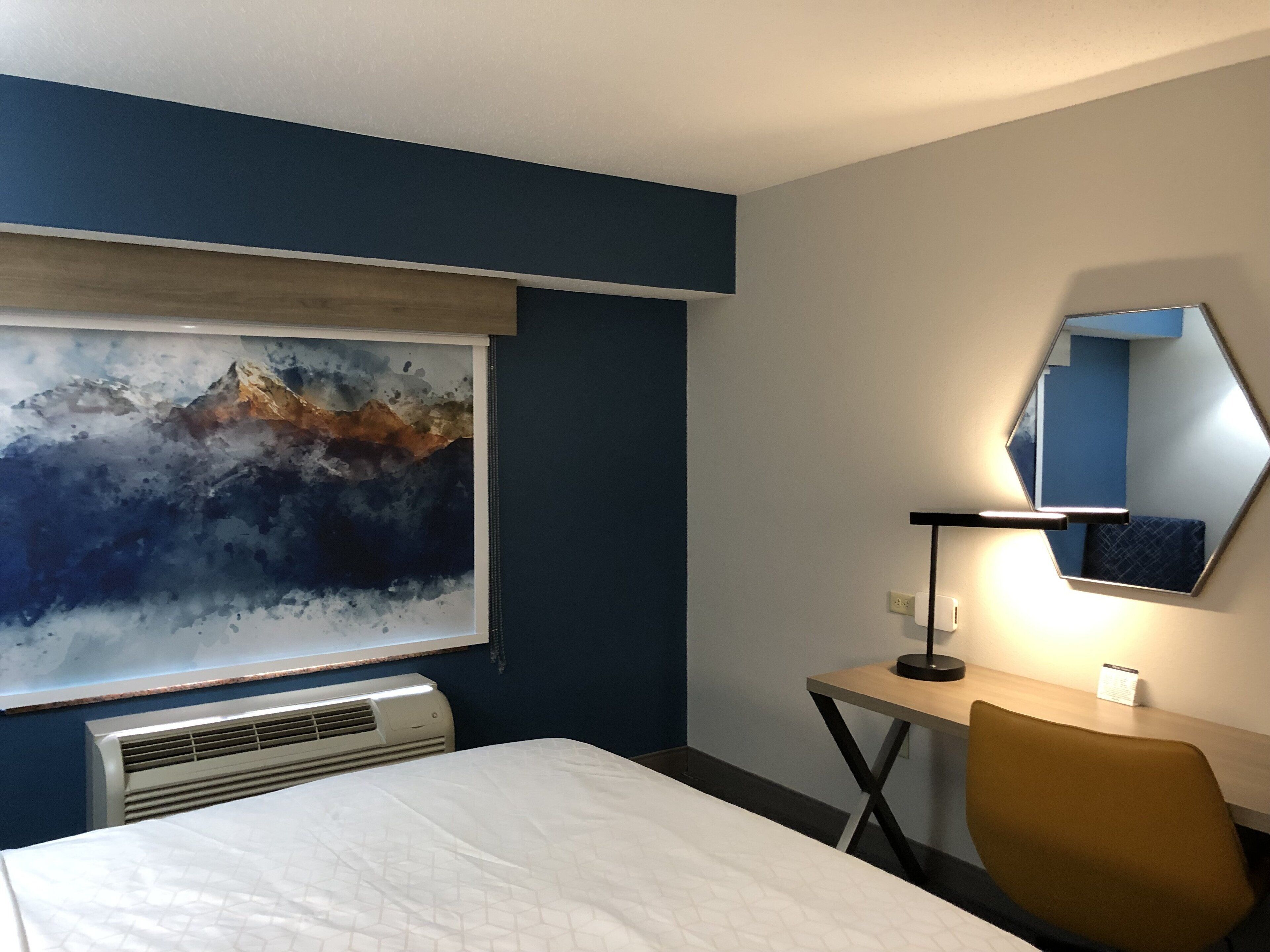 A hotel room with a bed, a desk, a chair, a large abstract painting, a mirror, and an air conditioning unit.