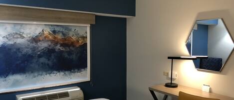 A hotel room with a bed, a desk, a chair, a large abstract painting, a mirror, and an air conditioning unit.