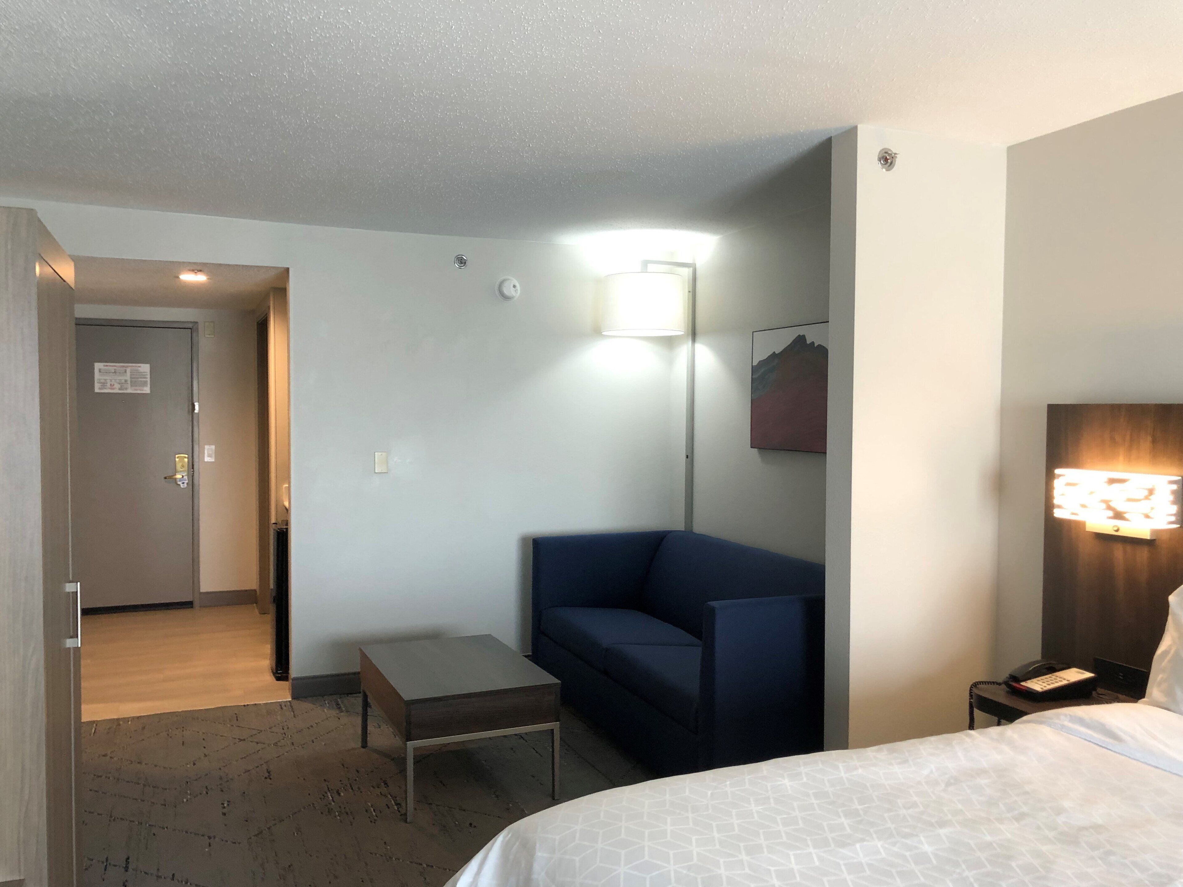 A hotel room with a bed, a small desk, a chair, a lamp, and a door leading to another room.