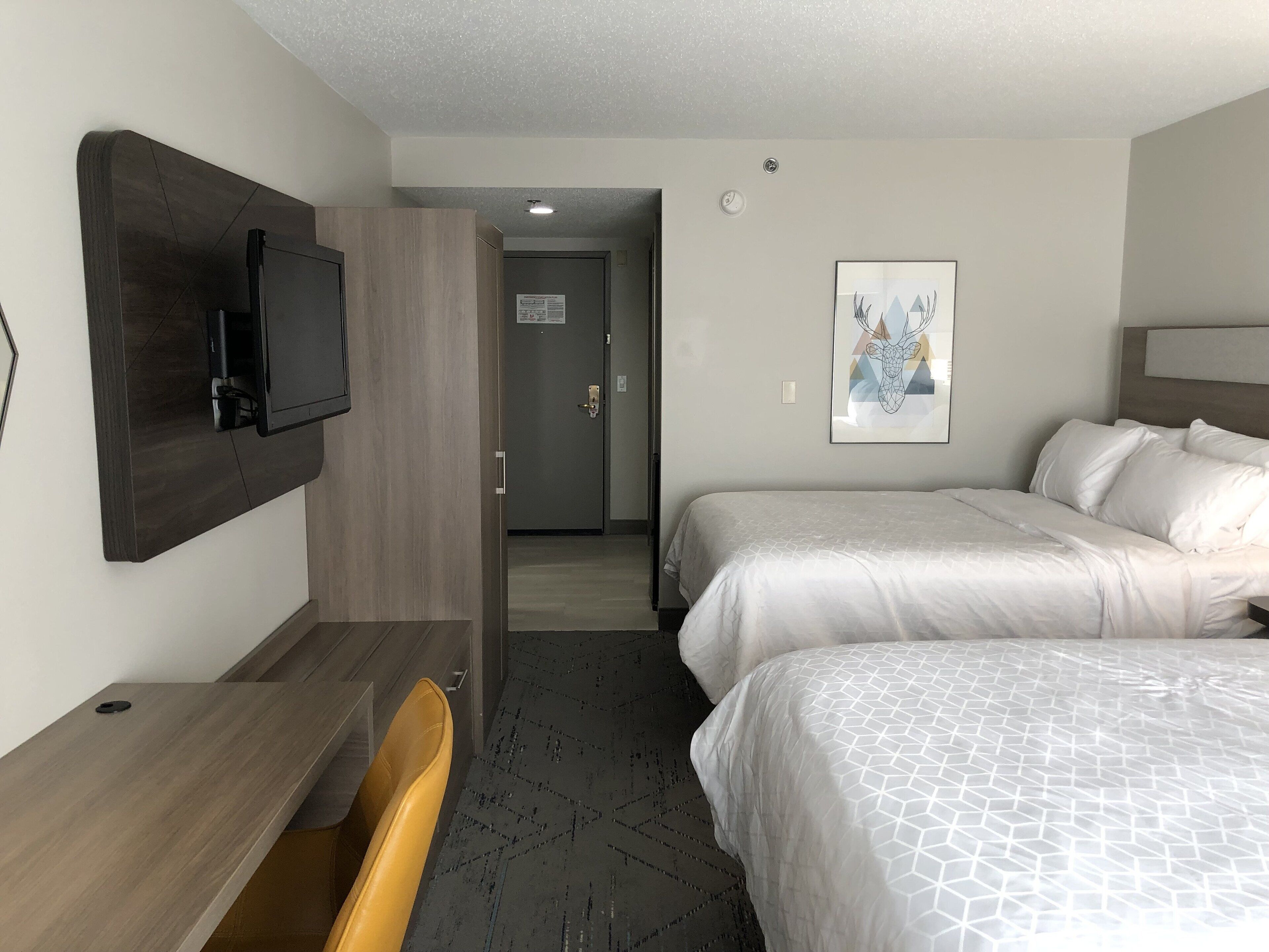 A hotel room with two beds, a desk, a chair, a TV mounted on the wall, and a door leading to another room.