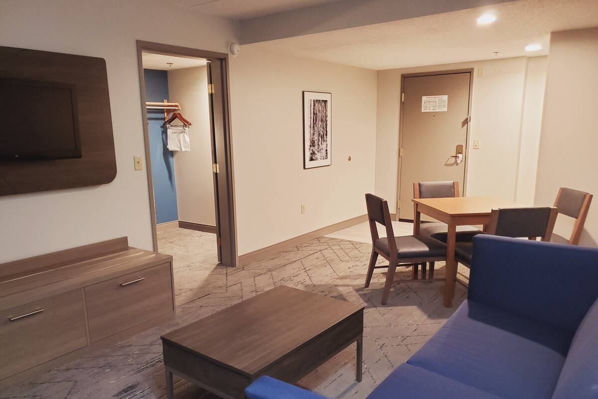 A room with a sofa, a wooden table, a TV mounted on the wall, and a door leading to another room.