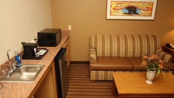 A compact kitchenette with a microwave, sink, and a small refrigerator. There is a striped sofa and a wooden table with a vase of flowers. A framed picture of a bear is on the wall.