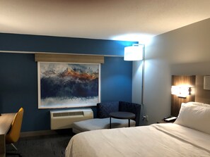 A hotel room with a bed, a small table, a chair, a painting on the wall, and a lamp.