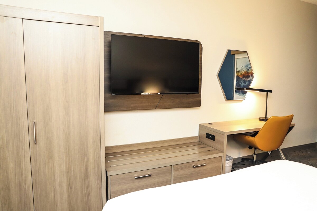 A hotel room with a bed, a desk with a chair, a wardrobe, a flat-screen TV, and a mirror.