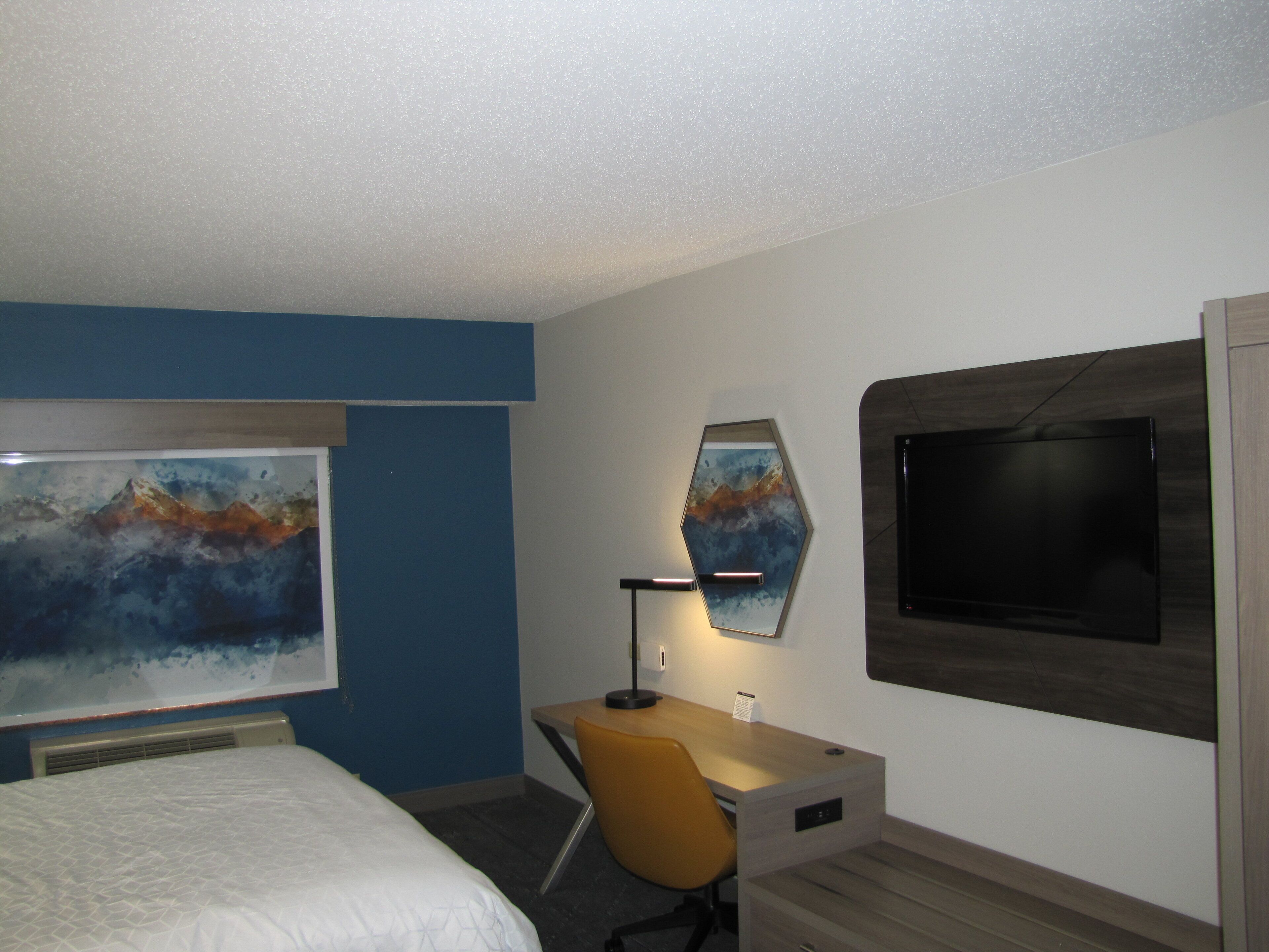 Ein Hotelzimmer mit Bett, Schreibtisch, Sessel, Flachbildfernseher und einem großen abstrakten Gemälde an der Wand.