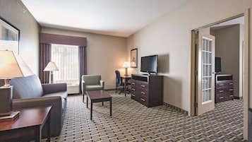A hotel room with a sofa, armchair, desk, TV, and a patterned carpet.