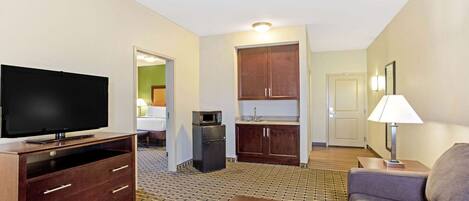 A hotel room with a flat-screen TV, a wooden dresser, a kitchenette, a sofa, and a patterned carpet.