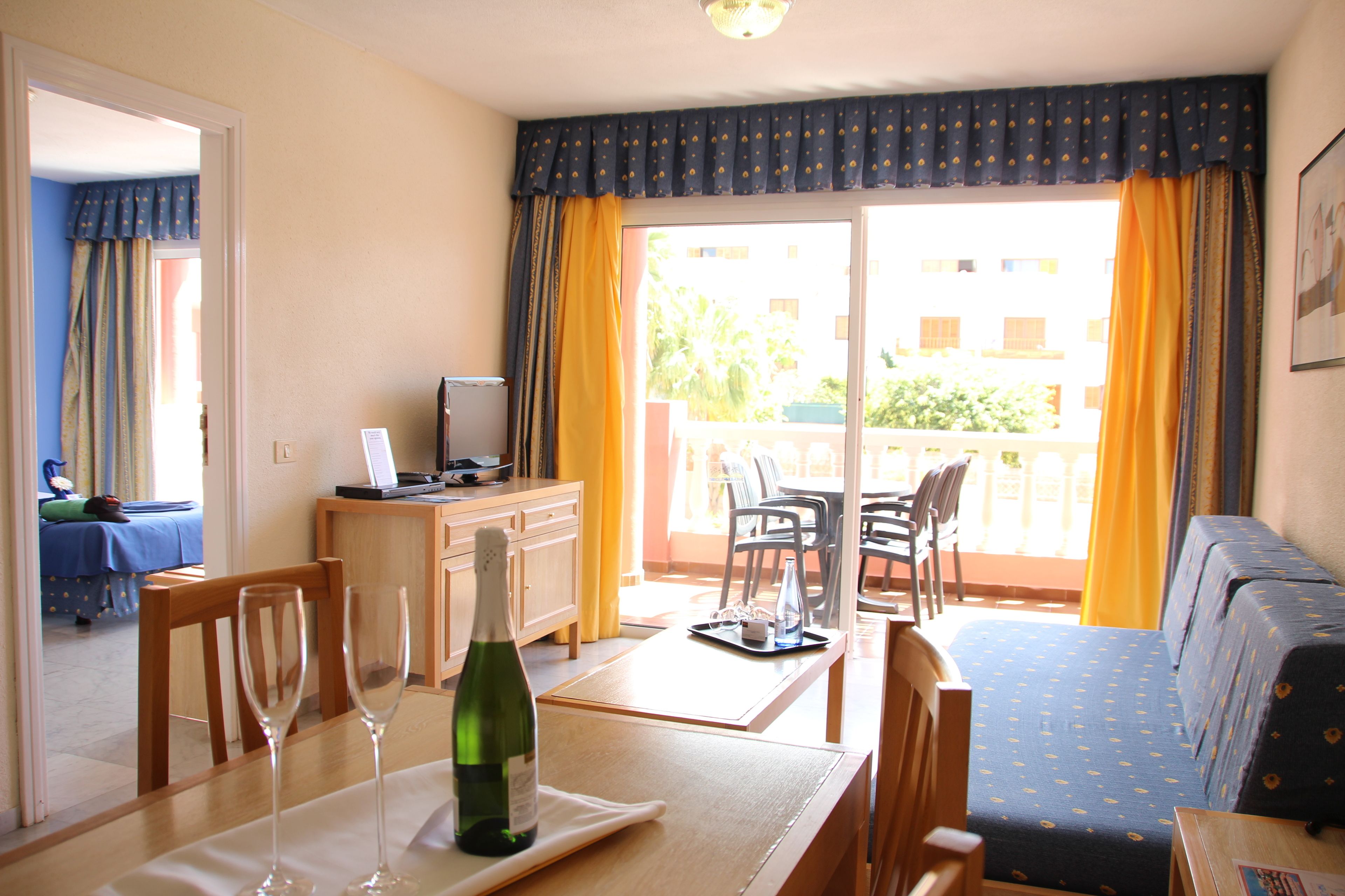 A room with a dining table, two wine glasses, a bottle of champagne, a sofa, and a balcony with yellow curtains.