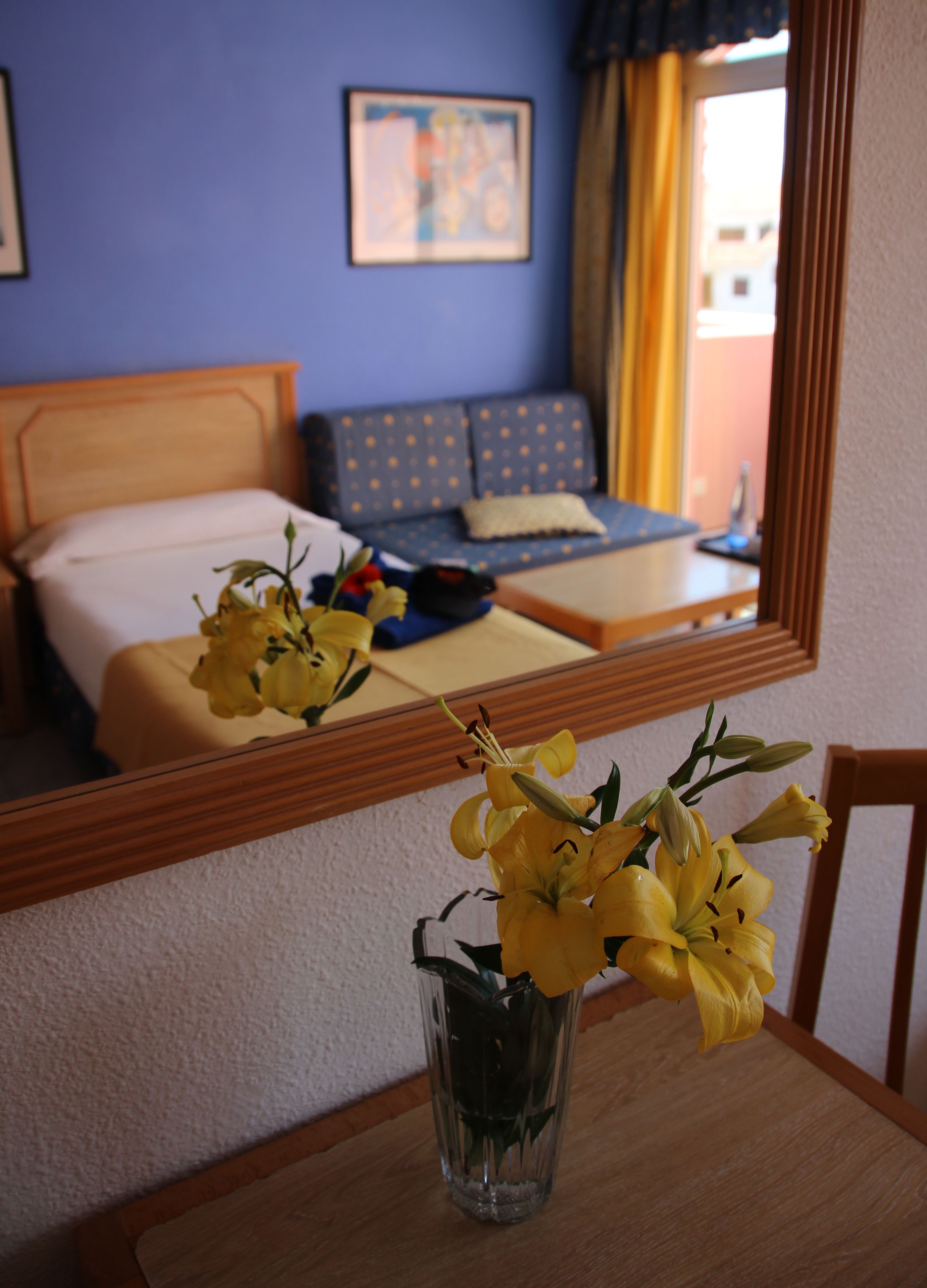 A hotel room with a bed, a sofa, a table with a vase of yellow flowers, and a mirror reflecting the room.