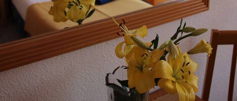 A hotel room with a bed, a sofa, a table with a vase of yellow flowers, and a mirror reflecting the room.