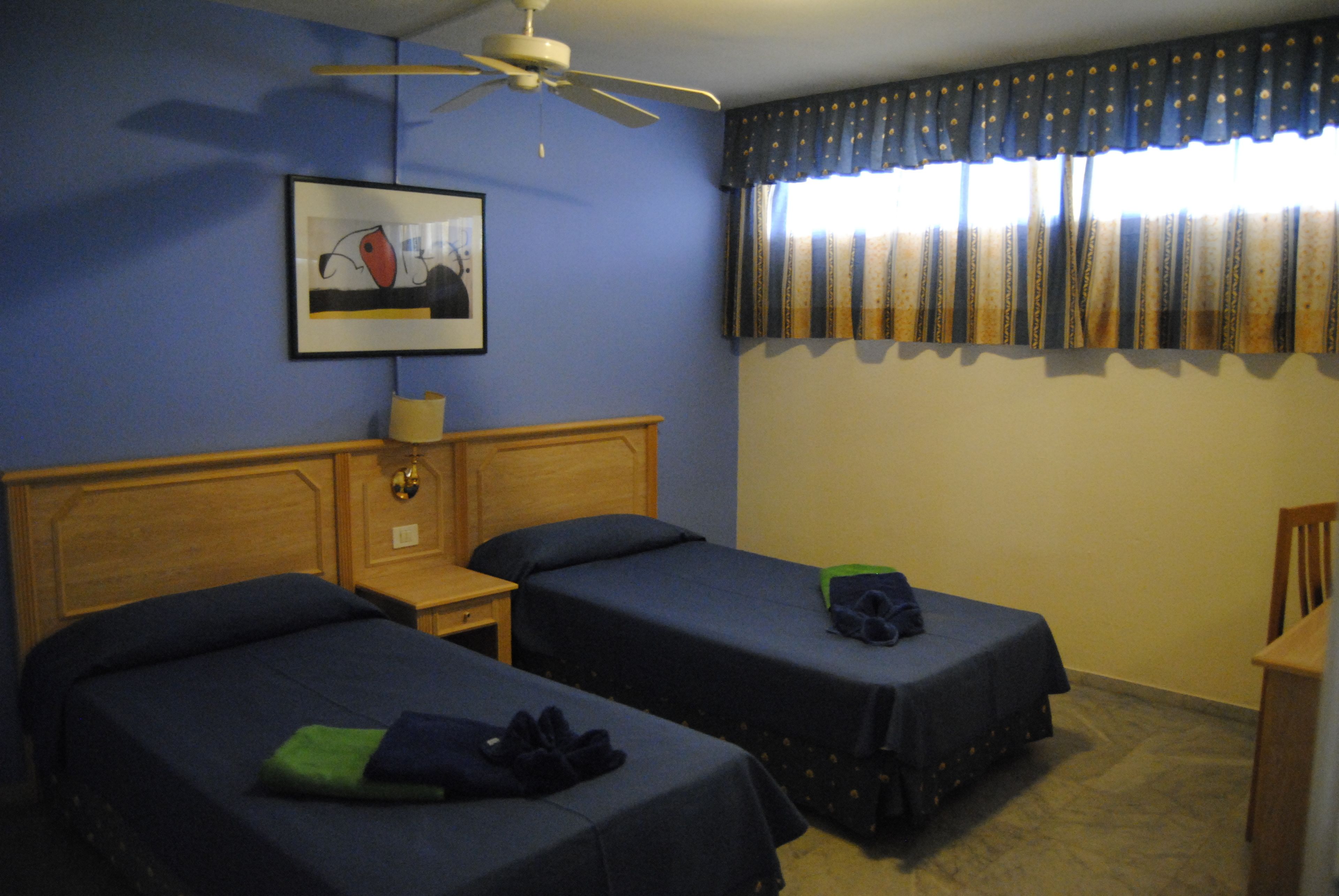 A hotel room with two single beds, a wooden headboard, a ceiling fan, a framed picture on the wall, and a window with curtains.