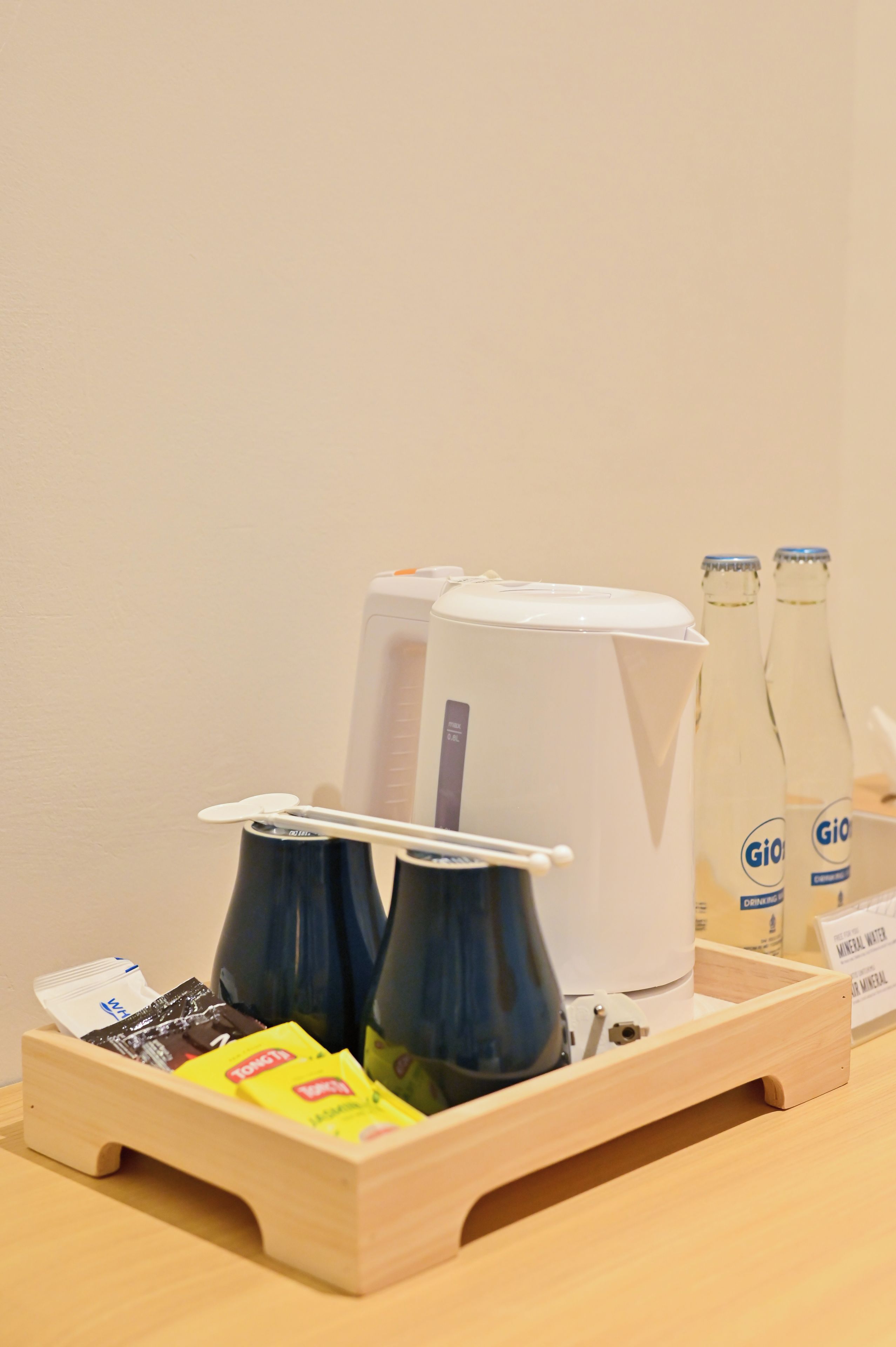 A wooden tray with a coffee machine, two cups, a bottle of water, and packets of snacks.