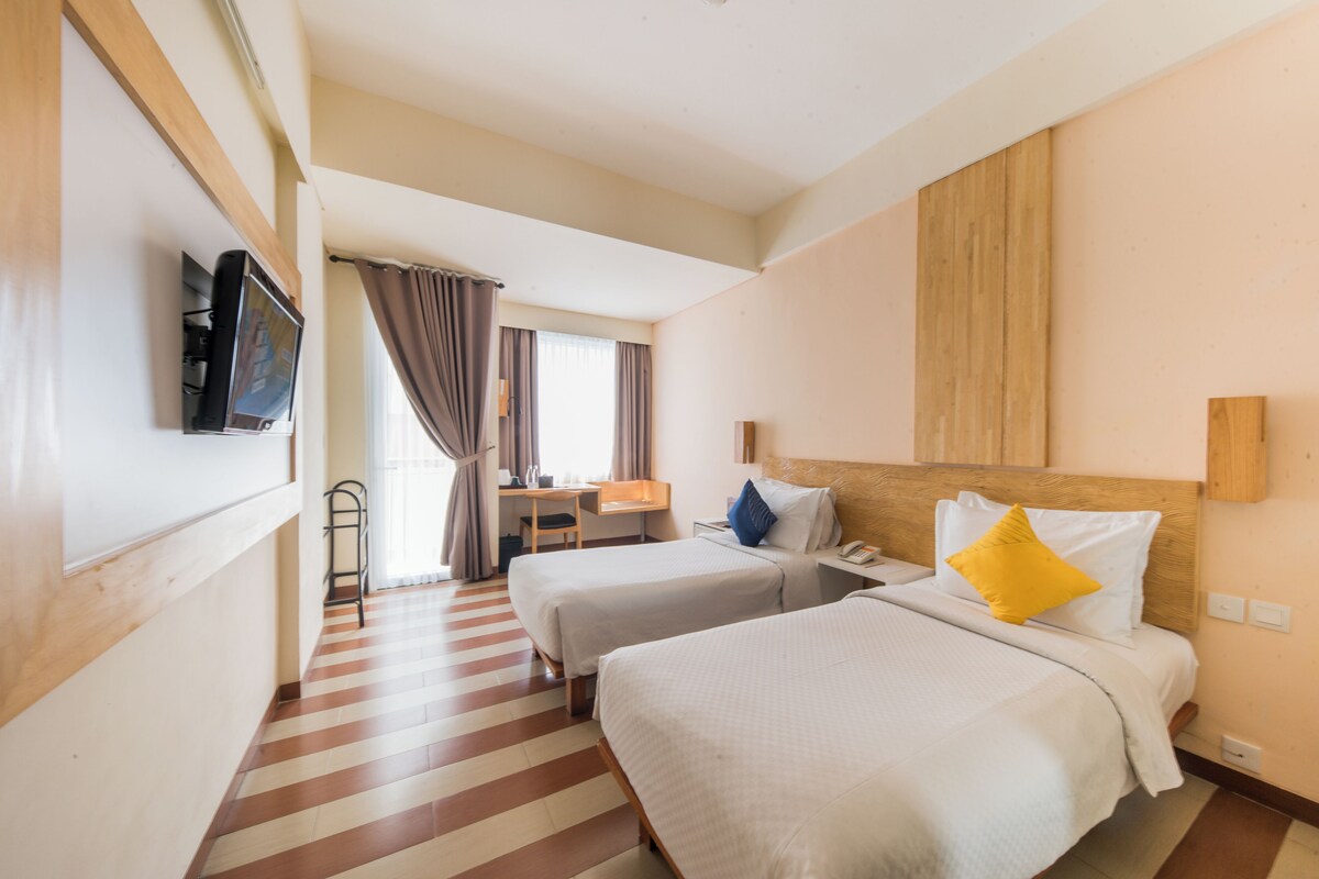 A hotel room with two beds, a desk, a chair, a TV, and a window with curtains.