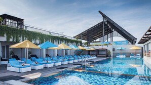 A swimming pool with lounge chairs and umbrellas, surrounded by a modern building with a sloped roof and a balcony with greenery.