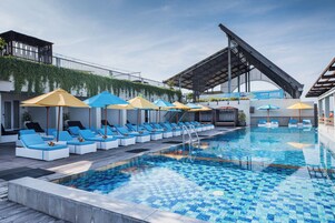 A swimming pool with lounge chairs and umbrellas, surrounded by a modern building with a sloped roof and a balcony with greenery.