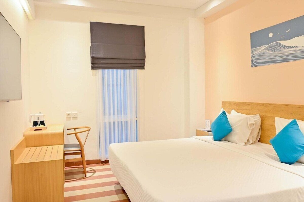 A hotel room with a bed, a desk, a chair, a window with curtains, and a wall painting.