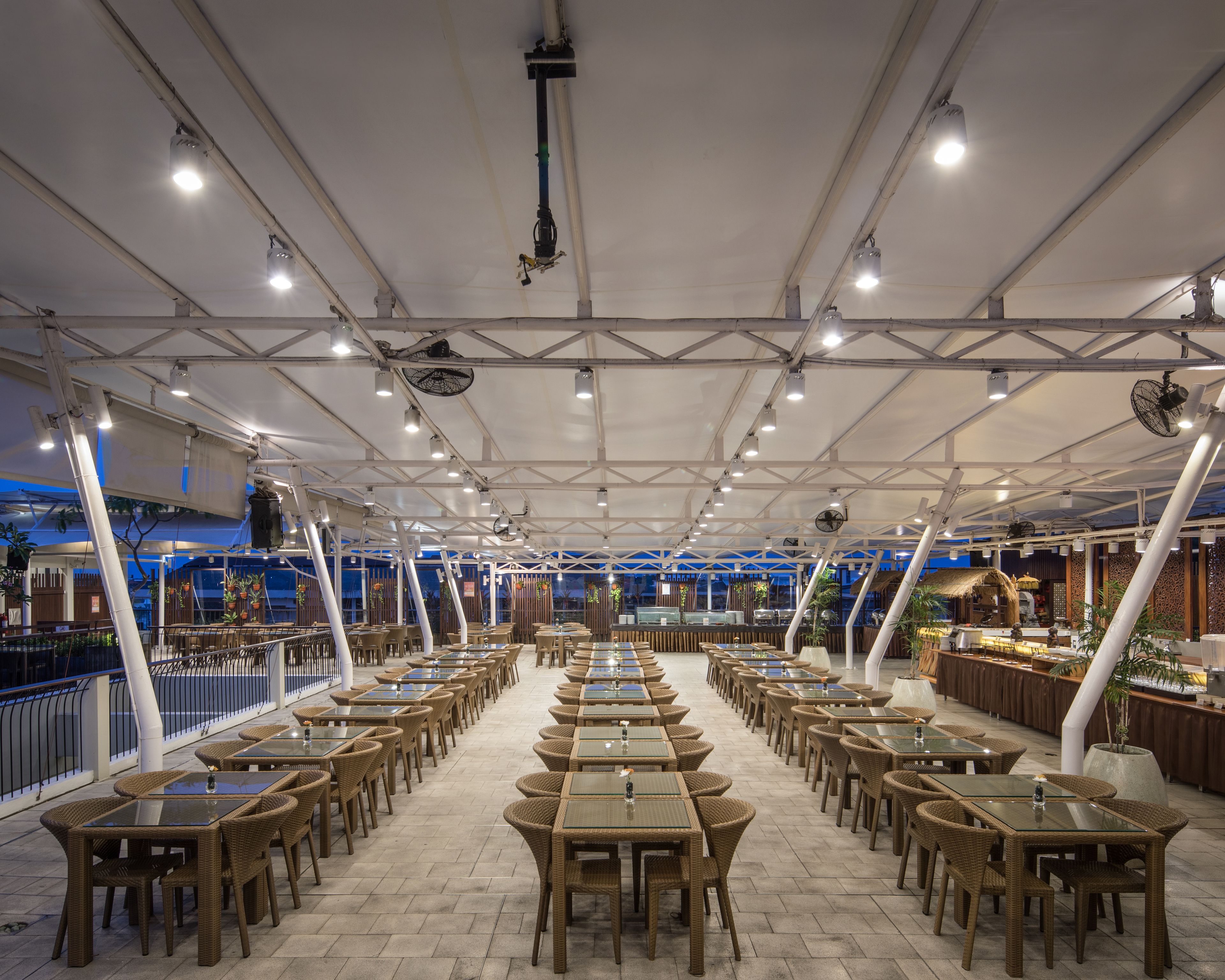 A spacious outdoor dining area with multiple tables and chairs, a covered ceiling, and a view of the cityscape in the background.