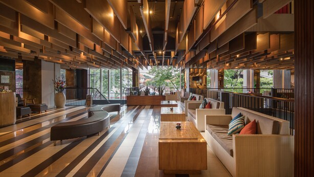 A spacious lobby with a modern design, featuring a central seating area with sofas and a coffee table, surrounded by a long corridor with a striped floor pattern.