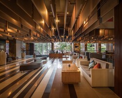A spacious lobby with a modern design, featuring a central seating area with sofas and a coffee table, surrounded by a long corridor with a striped floor pattern.