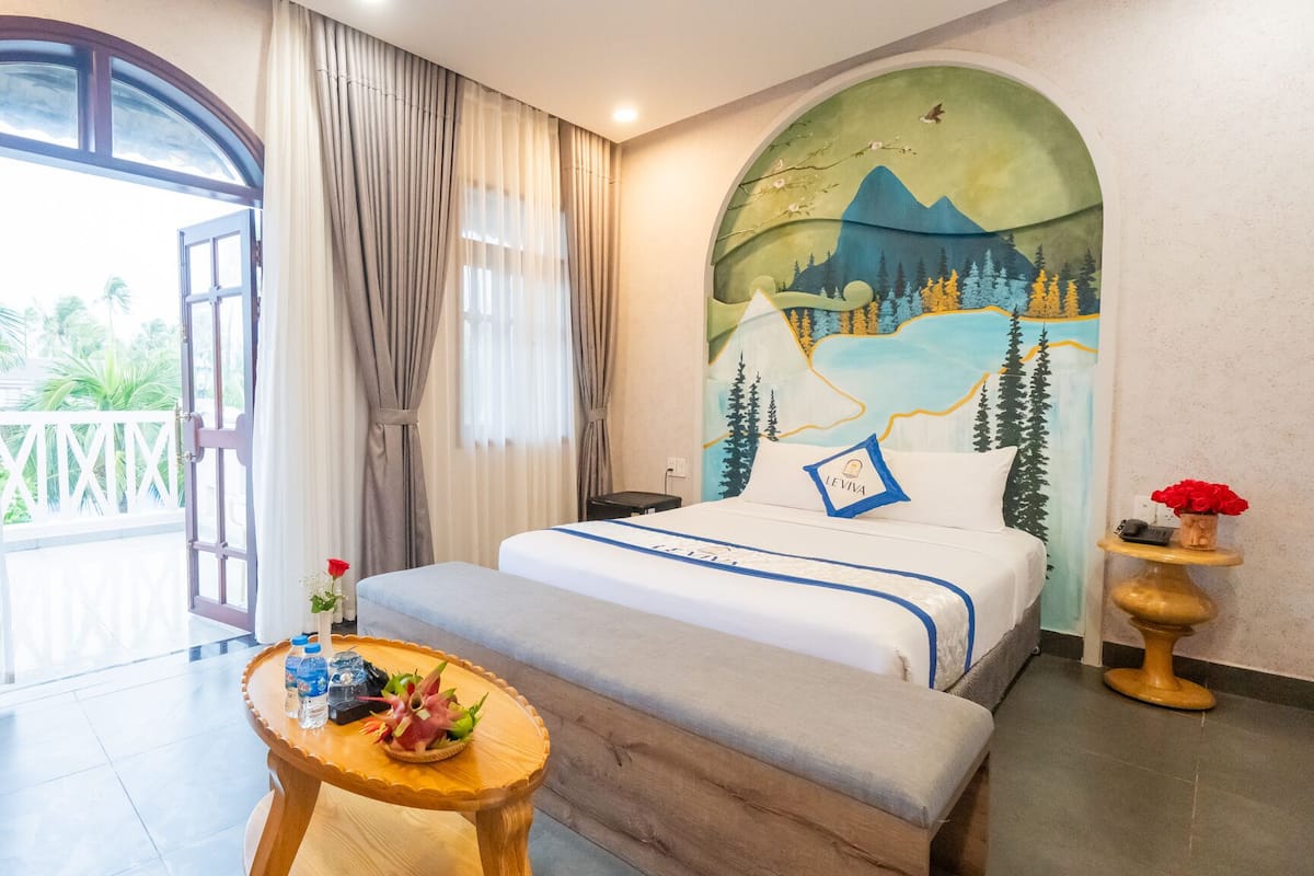 A hotel room with a bed, a small table with water bottles and snacks, a view of greenery outside, and a mural on the wall.