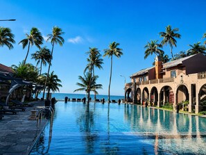 A resort pool with clear blue water, surrounded by palm trees and a building with arches. - Le Viva Mui Ne Resort (Mui Ne)
