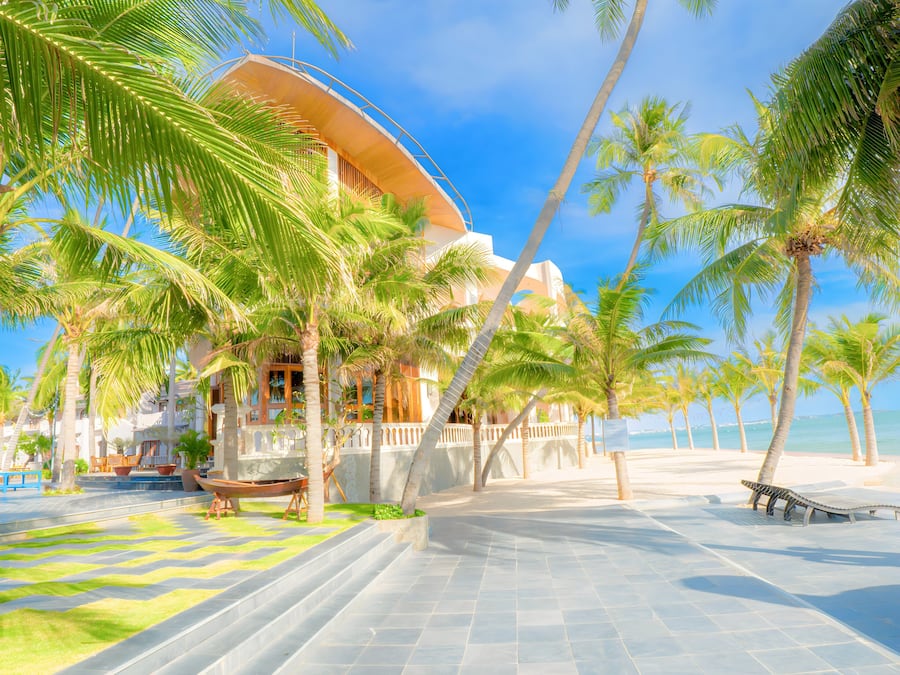 Um hotel à beira-mar com fachada curva, palmeiras e um calçadão de azulejos que leva à praia.
