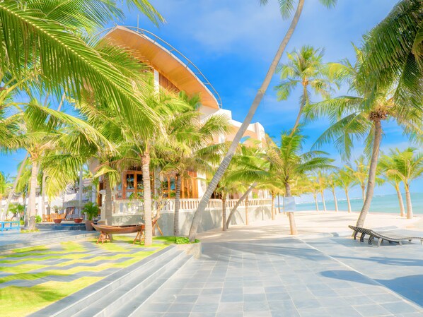 Un hotel frente al mar con fachada curva, palmeras y un camino empedrado que lleva a la playa.