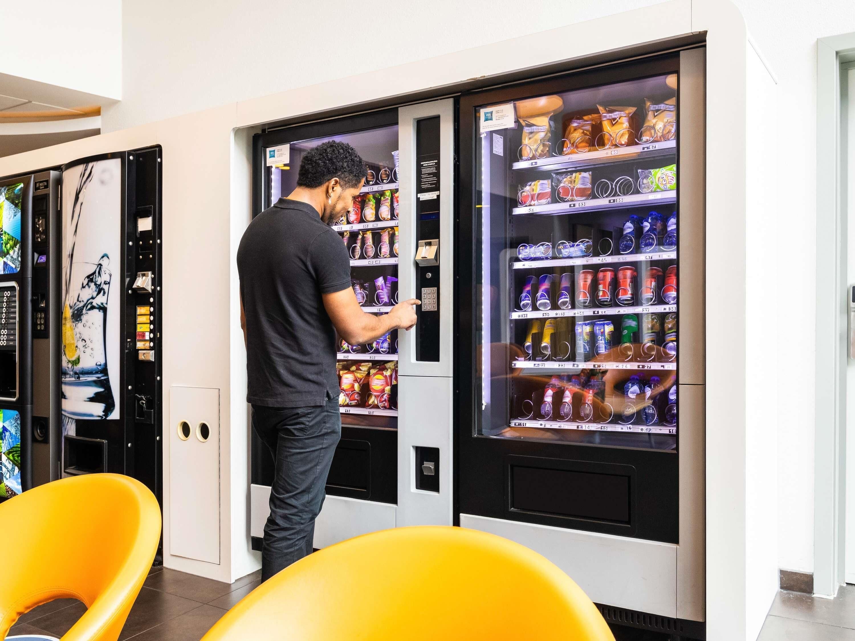 A person interacting with a vending machine in a modern setting with yellow chairs and a wall-mounted artwork.