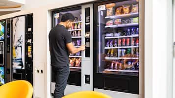 A person interacting with a vending machine in a modern setting with yellow chairs and a wall-mounted artwork.