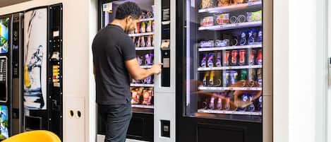 Eine Person bedient einen Verkaufsautomaten in einem modernen Ambiente mit gelben Stühlen und einem Wandbild.