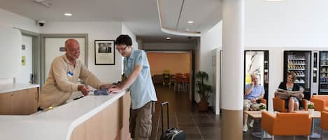 A reception area with a counter, a security camera, a framed photo, and a seating area with a plant and a vending machine.