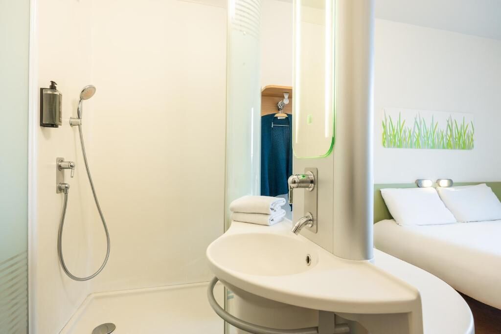 Un baño con ducha, lavabo y cama con pared verde y ropa de cama blanca.