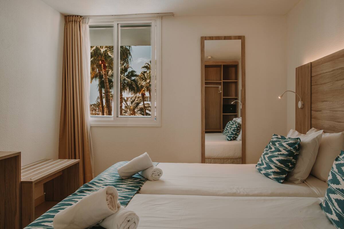 Ein Hotelzimmer mit zwei Betten, einem Holzkopfende, einem Spiegel und einem Fenster mit Vorhängen.