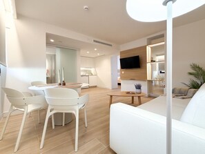Uma sala de estar moderna com um sofá branco, uma mesa de jantar com cadeiras e uma TV fixada na parede.