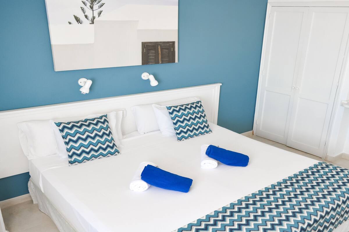 Ein ordentlich bezogenes Bett mit weißen Leinentextilien, blau-weiß gemusterten Kissen und einer blauen Decke.