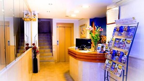 A hotel reception area with a curved reception desk, a staircase, a flower arrangement, and a magazine rack. - Hotel Sevilla (Almeria)