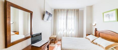 Habitación de hotel con una cama de madera, un escritorio con una computadora portátil, un espejo, un televisor y una ventana con cortinas.